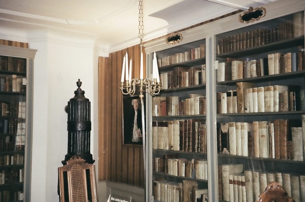 A corner of a room with a chandeliere, radiator, chair and painting floating amongst tall bookcases full of old, worn books.